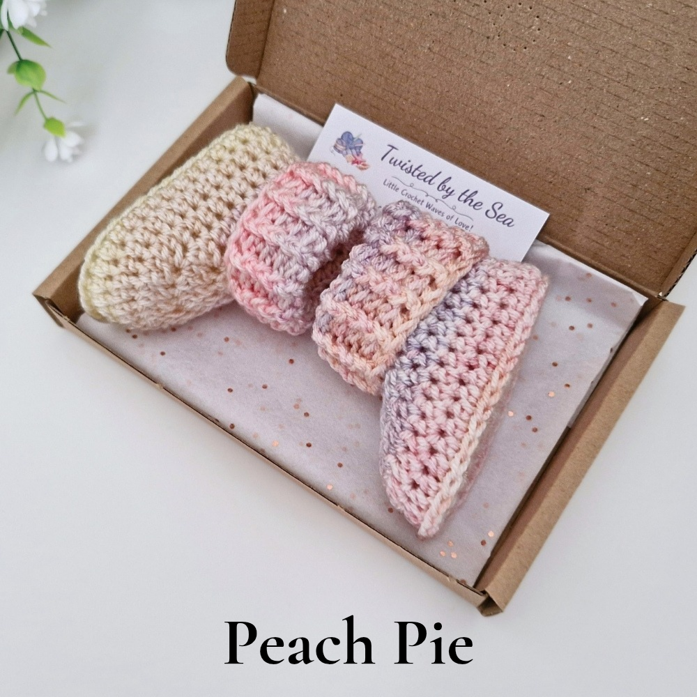 Two crocheted baby booties in a soft peach, pink, and lavender variegated yarn, presented in an open brown cardboard gift box with a branded card. The text 'Peach Pie' is displayed at the bottom of the image.