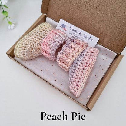 Two crocheted baby booties in a soft peach, pink, and lavender variegated yarn, presented in an open brown cardboard gift box with a branded card. The text 'Peach Pie' is displayed at the bottom of the image.