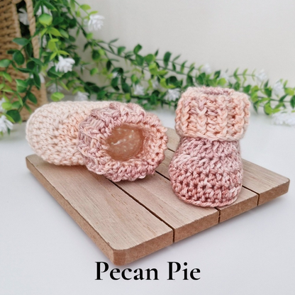 Two hand-crocheted baby booties in soft pastel shades of pink, peach, and cream, displayed on a small wooden block with the text 'Pecan Pie' underneath.