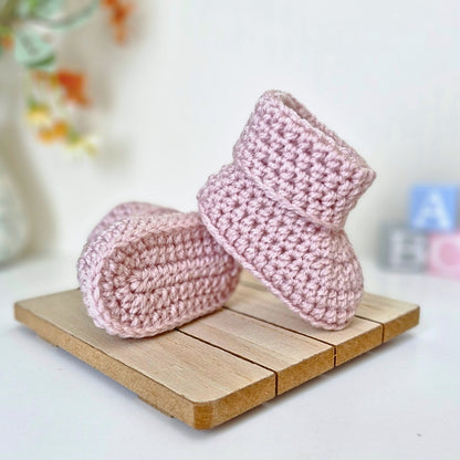 Pink crocheted baby booties on a wooden coaster with a blurred background