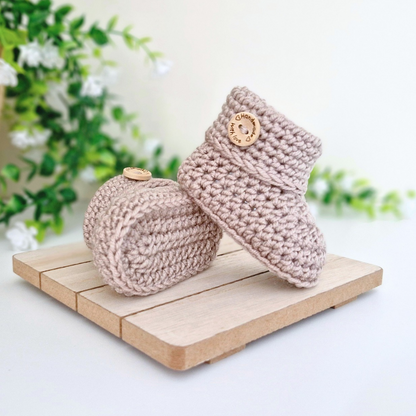 A pair of crochet baby booties in a light taupe-beige color, displayed on a small wooden block with a soft-focus background of green leaves and white flowers.