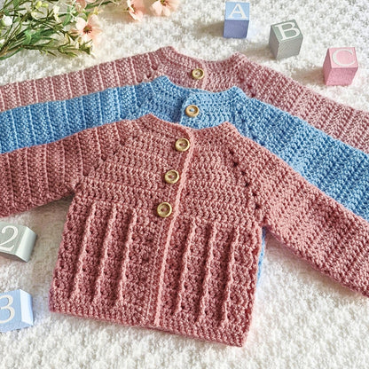 Three crocheted baby cardigans, two pink and one blue, on a textured surface.