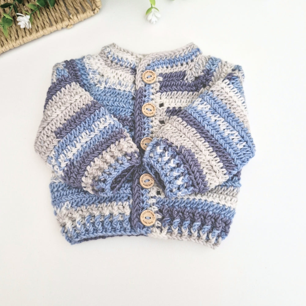 A handmade crocheted baby cardigan with horizontal stripes in light blue, medium blue, and cream, featuring wooden buttons down the front, laid flat on a white surface.