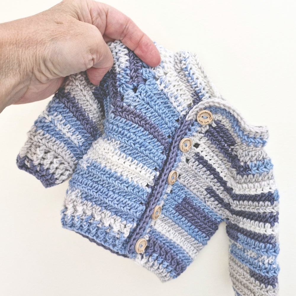 A close-up of a hand holding a small crocheted cardigan with horizontal stripes in light blue, medium blue, navy, and white. The cardigan has a button closure with light wood buttons, set against a plain off-white background.