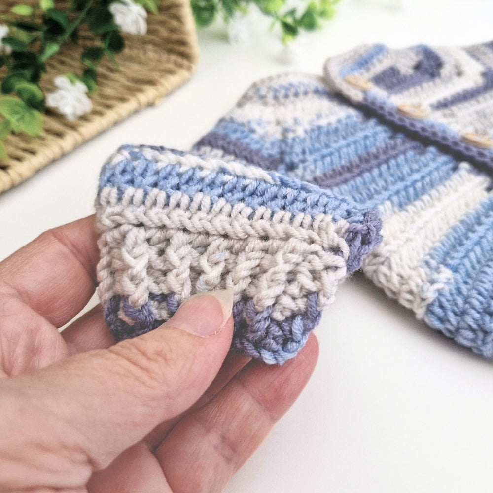 A close-up of a hand holding a small crocheted textile piece with blue, white, and gray stripes, with more matching crocheted items blurred in the background on a white surface.
