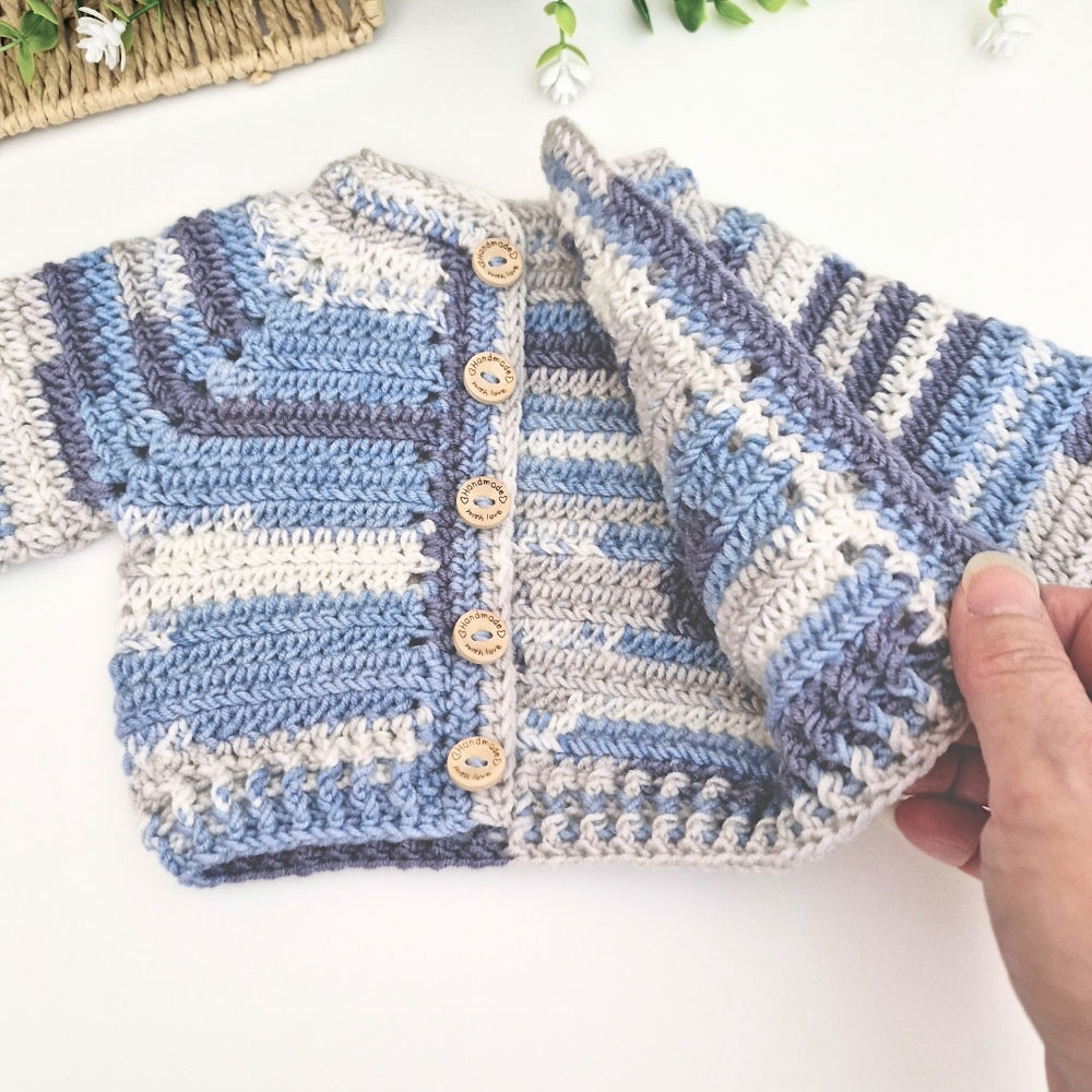 A handmade crocheted baby cardigan with blue, gray, and cream stripes, featuring wooden buttons, held open by a hand on a white background.