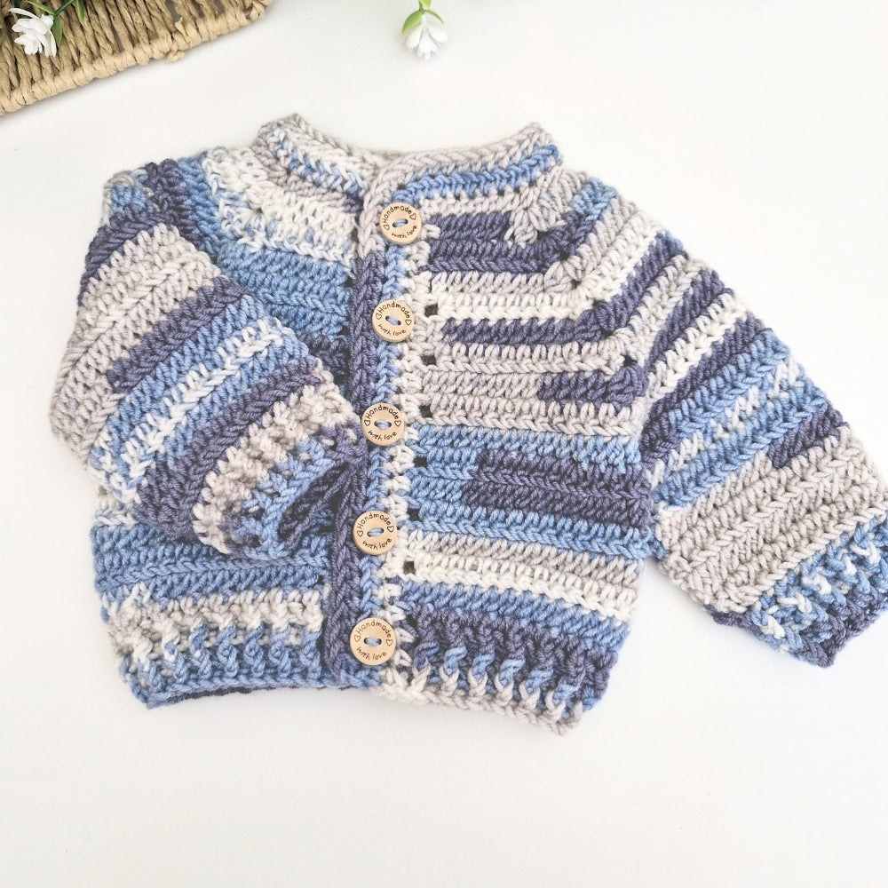 A handmade crocheted cardigan sweater for baby, featuring horizontal stripes in light blue, slate blue, gray, and cream, with four wooden buttons on the front. It is laid flat on a white surface.
