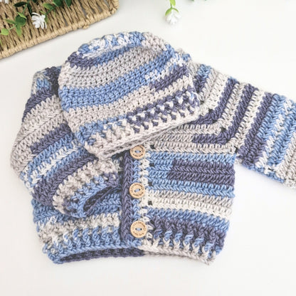 A handmade crocheted baby cardigan and matching hat set in blue, gray, and white stripes, styled on a white background with a woven mat and small white flowers.