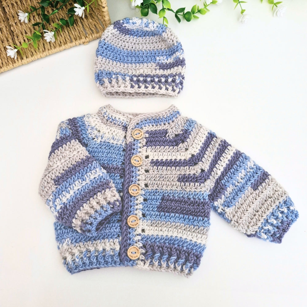 A handmade crocheted baby outfit set featuring a striped cardigan and matching beanie hat, laid flat on a white background with decorative green leaves and a woven basket in the corner.