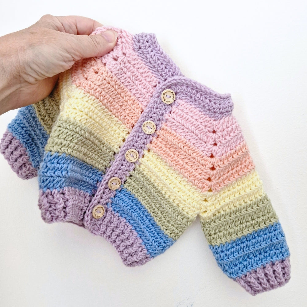 A hand holds a small handmade crocheted cardigan with pastel rainbow stripes in pink, yellow, blue, green, and lavender, featuring wooden buttons on a white background.