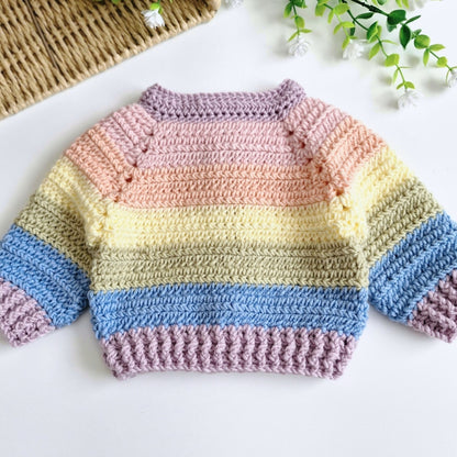 A handmade, crocheted baby sweater with horizontal pastel stripes in pink, yellow, green, and blue, laid flat on a white surface with decorative props.