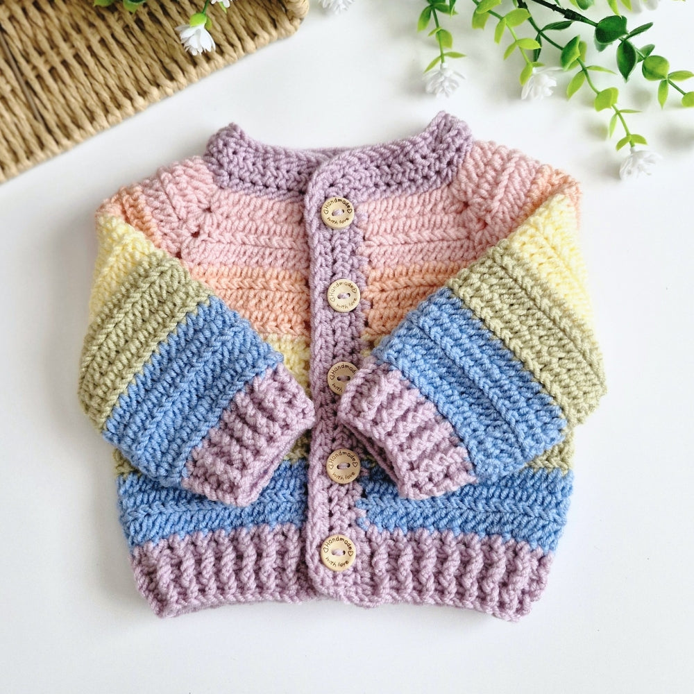 A handmade crocheted baby cardigan with pastel rainbow stripes in pink, orange, yellow, blue, and lavender, featuring wooden buttons, laid flat on a white surface with decorative props.