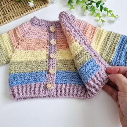 A handmade crocheted baby cardigan with pastel rainbow stripes in pink, yellow, green, blue, and lavender, featuring wooden buttons down the front. A hand is holding up the sleeve of the cardigan, and a small decorative plant and woven basket are visible in the background.