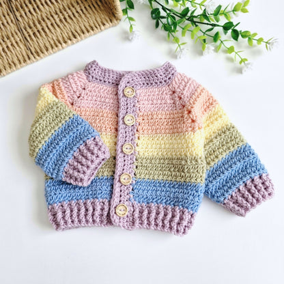 A handmade crocheted rainbow baby cardigan sweater in muted pastel rainbow stripes in pink, yellow, blue, and lavender, featuring four wooden buttons, laid flat on a white surface with decorative props.