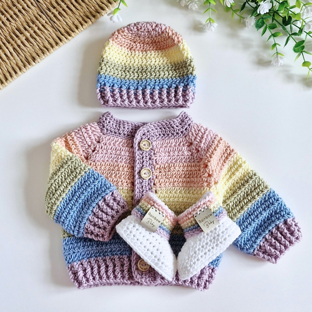 A handmade crochet rainbow baby outfit set featuring a striped cardigan, matching beanie, and booties, laid flat on a white surface with decorative props.