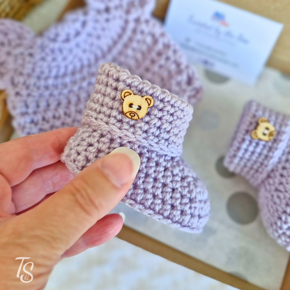 Hand holding lavender crochet baby bootie with wooden bear button, matching bootie in box