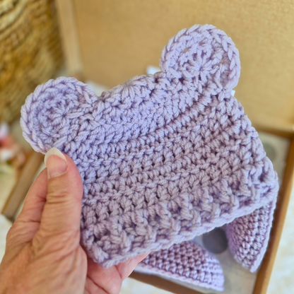 Hand holding a lilac shetland wool crochet baby hat with teddy bear ears in a box