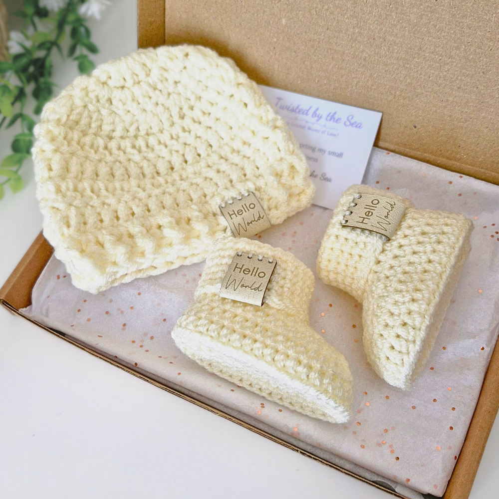 Set of cream crocheted baby booties and hat with 'Hello World' tags in a box.