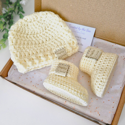 Set of cream crocheted baby booties and hat with 'Hello World' tags in a box.