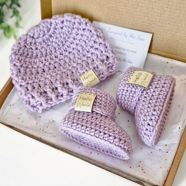 Set of purple baby booties and hat with 'Hello World' tags in a box.