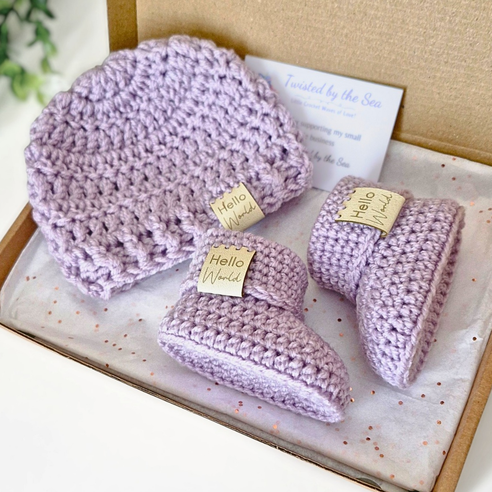 Set of purple baby booties and hat with 'Hello World' tags in a box.