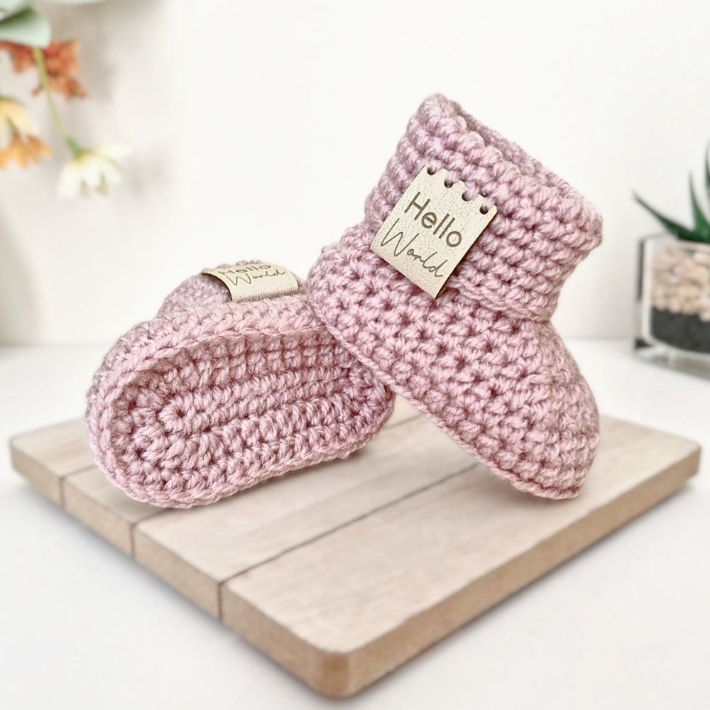 Pair of pink crochet baby booties with 'Hello World' tags on a wooden board.