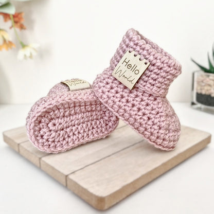 Pair of pink crochet baby booties with 'Hello World' tags on a wooden board.