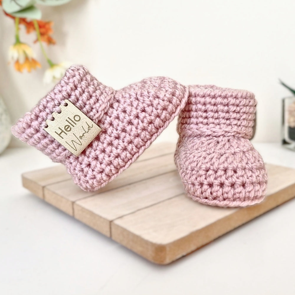 Pair of pink crochet baby booties with 'hello world' leather tags on a wooden surface with a blurred floral background.
