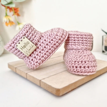 Pair of pink crochet baby booties with 'hello world' leather tags on a wooden surface with a blurred floral background.