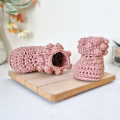 A pair of pink crochet baby booties on a wooden board with a blurred floral background