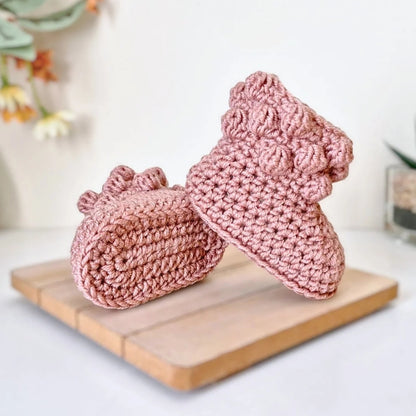 Pink crochet baby booties on a wooden board with a blurred floral background