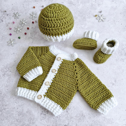 A crocheted baby mistletoe green set laid flat on a light textured surface, including an olive green and white cardigan with wooden buttons, a matching hat, and booties. Small decorative snowflake and bead ornaments are scattered around the items.