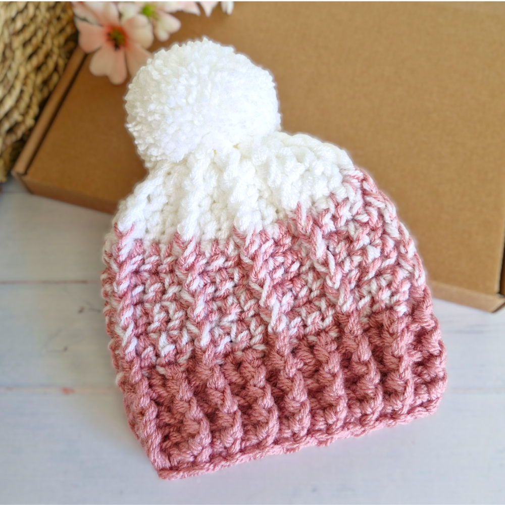 Pink and white crocheted beanie with a pom-pom on a light wooden surface.
