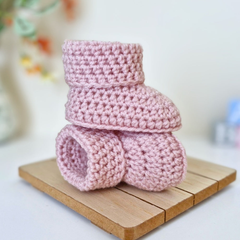 Two light pink crocheted baby booties stacked on a small wooden block, photographed against a softly blurred light background.
