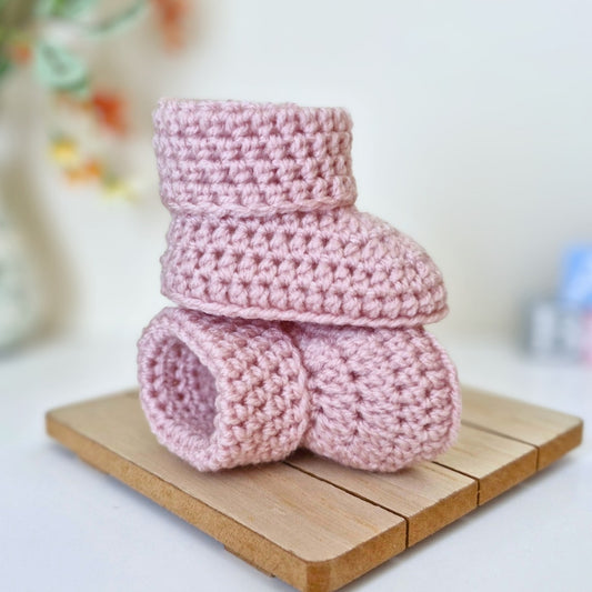 Two light pink crocheted baby booties stacked on a small wooden block, photographed against a softly blurred light background.