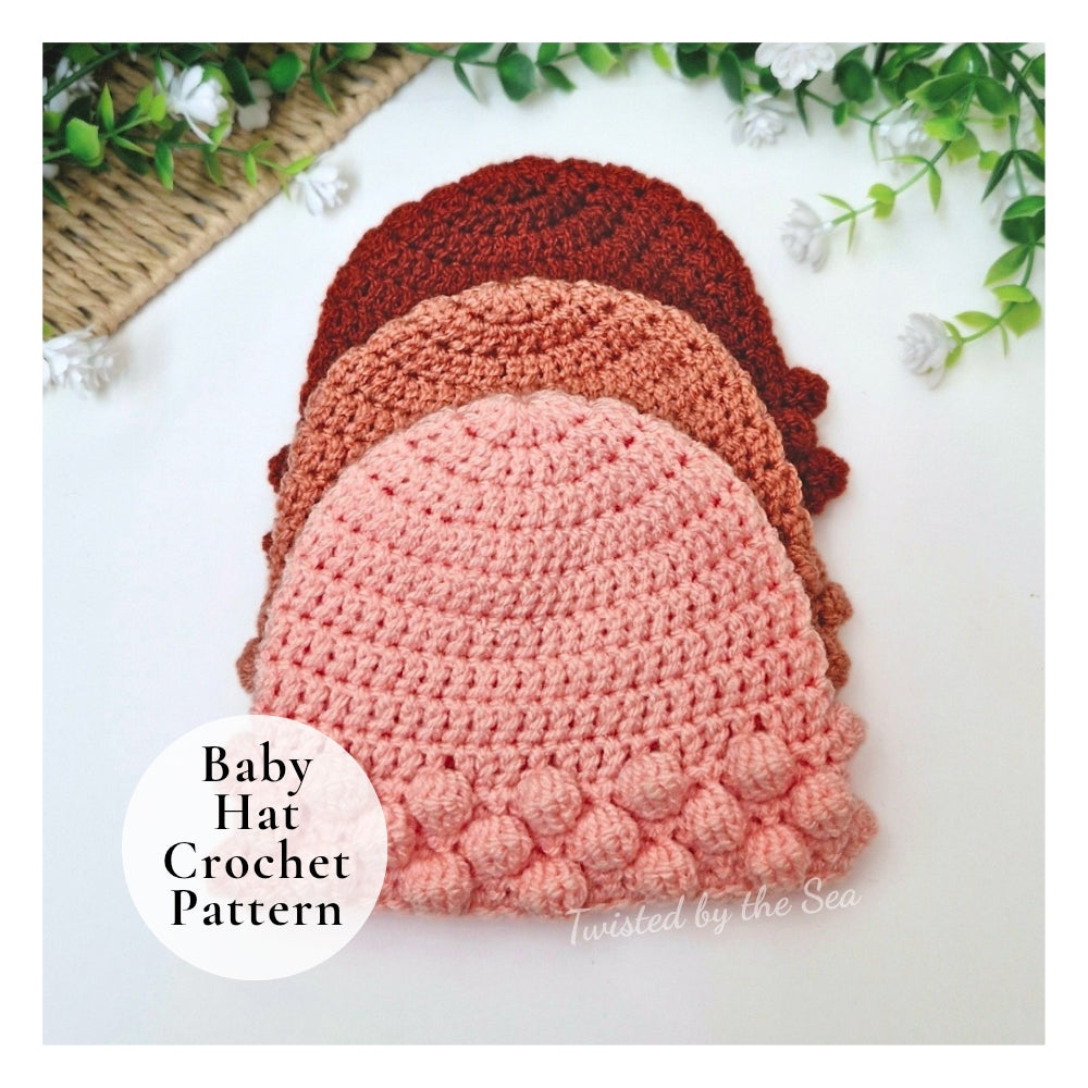 Three crocheted baby hats in stacked rows on a white background with decorative green leaves and white flowers. A white circular label reads 'Baby Hat Crochet Pattern' and 'Twisted by the Sea' appears near the bottom right.