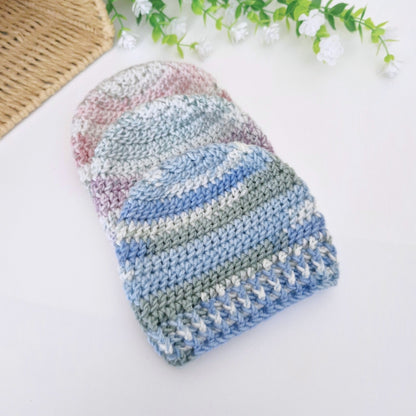 A handmade crocheted beanie hat with horizontal stripes of light blue, dusty blue, sage green, and soft pink, displayed on a clean white surface with decorative white flowers and a woven mat in the background.