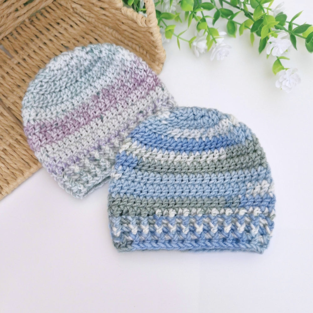 Two crocheted knit beanies in soft pastel stripes, one in blue, gray, and lavender, and the other in blue, gray, and white, displayed on a white surface with a wicker basket and green leaves as props.
