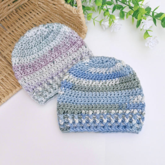 Two crocheted knit beanies in soft pastel stripes, one in blue, gray, and lavender, and the other in blue, gray, and white, displayed on a white surface with a wicker basket and green leaves as props.