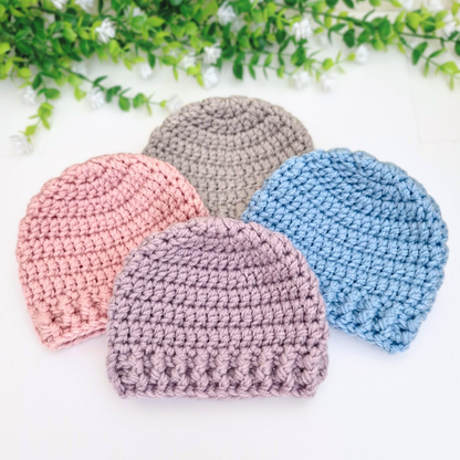 Four crocheted hats in purple, pink, grey, and blue on a white background with greenery.