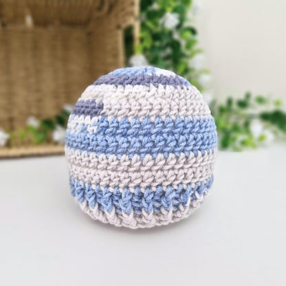 A handmade crocheted baby hat with horizontal stripes of light blue, off-white, and lavender purple, resting on a white surface with a blurred background.