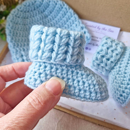 Blue knitted baby booties held by a hand with a matching hat in the background.