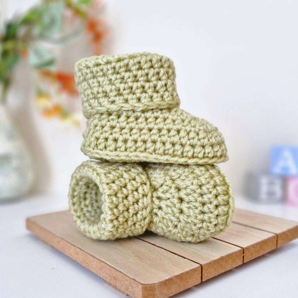 Two light sage green crocheted baby booties stacked on a small wooden block, with a softly blurred indoor background.