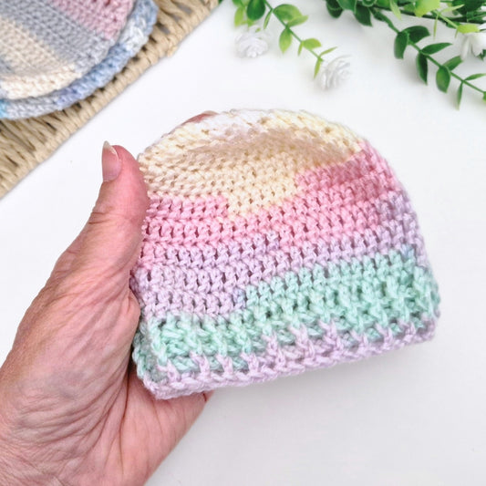 A hand holding a small crocheted beanie hat with pastel rainbow stripes in cream, yellow, pink, lavender, and mint green, with a second similar hat partially visible on a woven mat in the background.