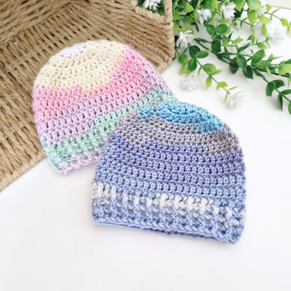 Two crocheted baby beanie hats displayed on a white surface with decorative props. One hat has pastel stripes in cream, pink, mint green, and lavender.