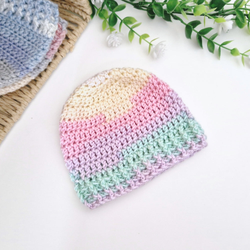 A pastel crocheted beanie hat with horizontal stripes of cream, pink, lavender, and mint green, displayed on a white surface with decorative greenery and another hat in the background.