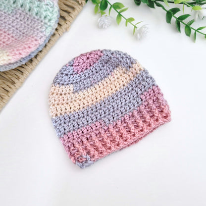 A handmade crocheted beanie hat with horizontal stripes of pink, lavender, cream, and light blue, displayed on a white surface with decorative props.