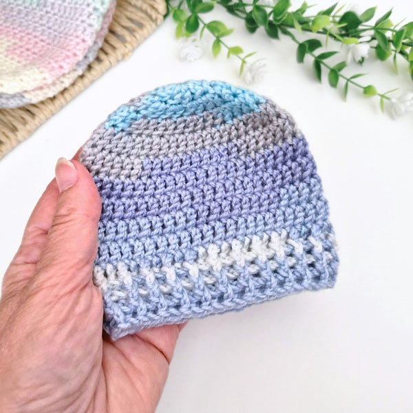 A hand holds a small crocheted beanie hat with horizontal stripes in light blue, lavender, gray, and white. The background is a clean white surface with decorative green leaves and a partially visible matching crocheted item.