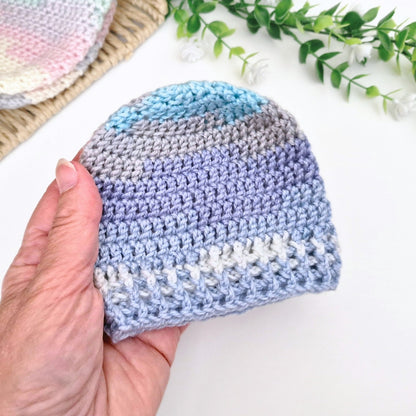 A hand holds a small crocheted beanie hat with horizontal stripes in light blue, lavender, gray, and white. The background is a clean white surface with decorative green leaves and a partially visible matching crocheted item.