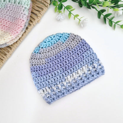 A crocheted baby beanie hat in pastel stripes of blue, purple, gray, and white, displayed on a white surface with decorative greenery and a woven mat.